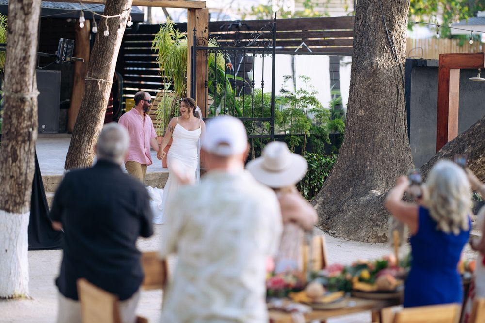 People gather outdoors at a garden venue with trees and wooden benches during a daytime event.