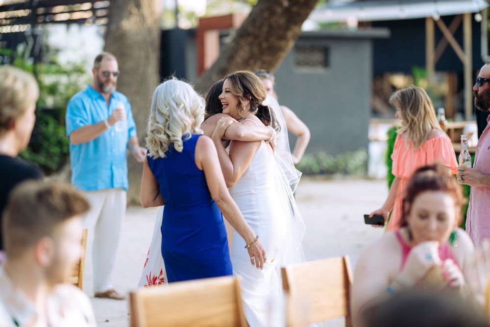 Guests mingle and embrace at an outdoor celebration with wooden benches and trees in the background.