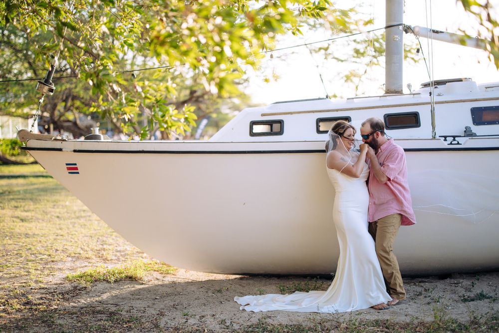 A couple shares a romantic moment beside a white sailboat on a sunny day with trees in the background.