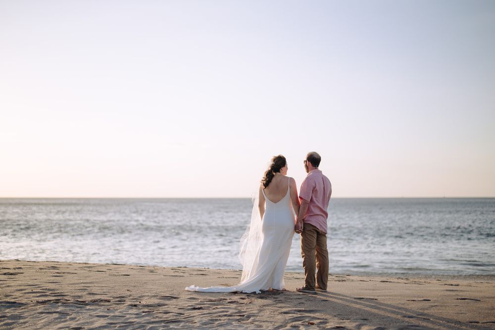 A couple stands together on a peaceful beach at sunset, the bride in white and her partner in pink.