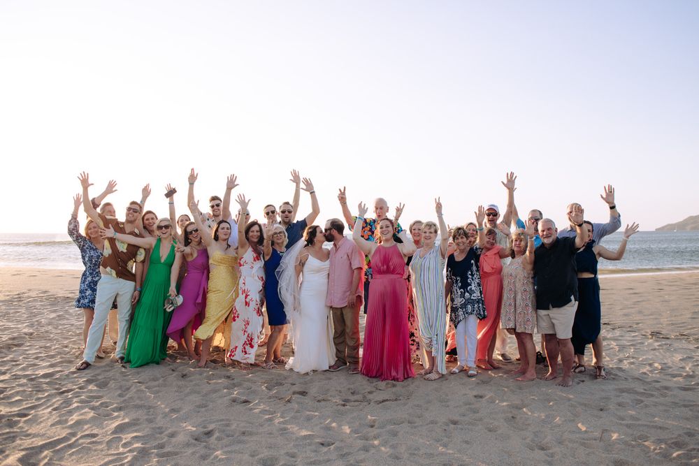 A large group celebrates joyfully on a beach at sunset with colorful dresses and festive energy.
