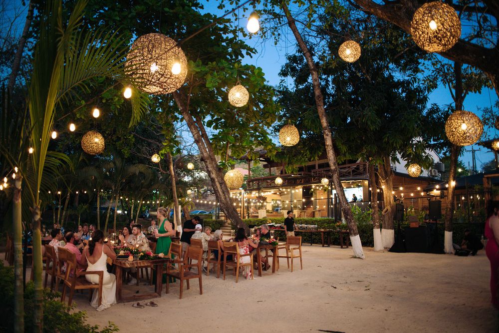 Outdoor evening wedding reception with illuminated wicker lanterns hanging over tables beneath tropical palm trees.