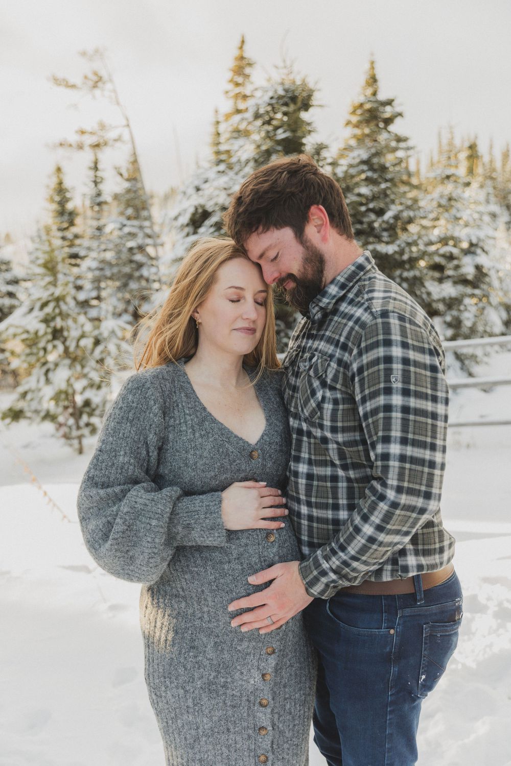 A couple embraces in a snowy winter forest setting wearing a grey sweater and plaid flannel shirt.