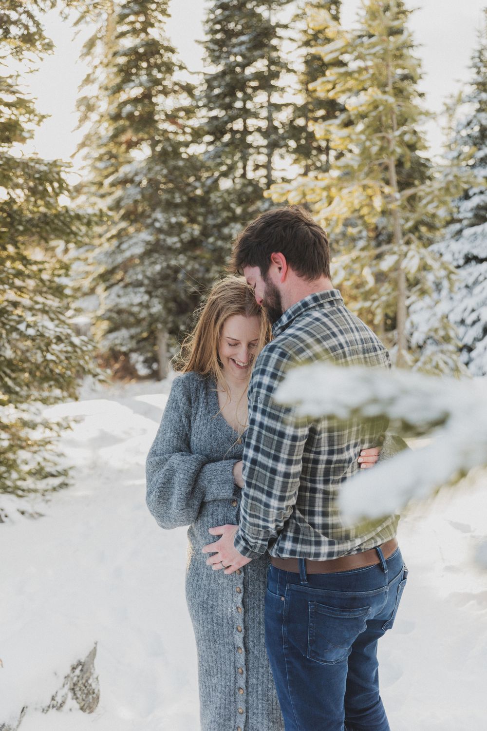 A couple shares an intimate embrace in a snowy winter forest setting with evergreen trees in the background.