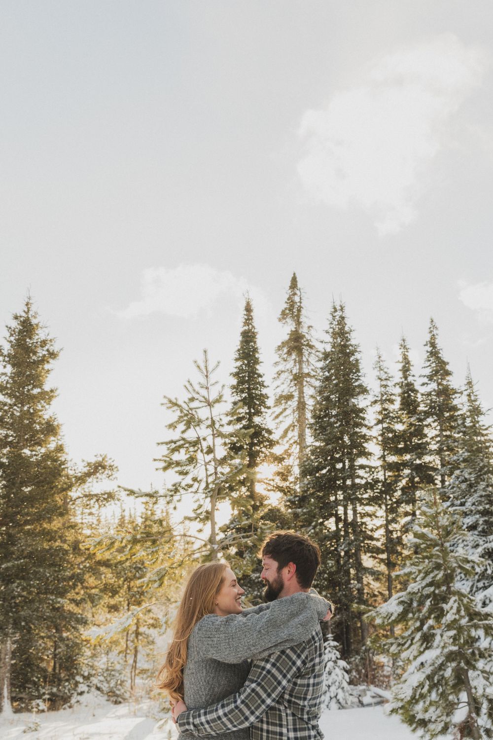 A couple embraces in a snowy winter forest with evergreen trees and sunlight peeking through the branches.
