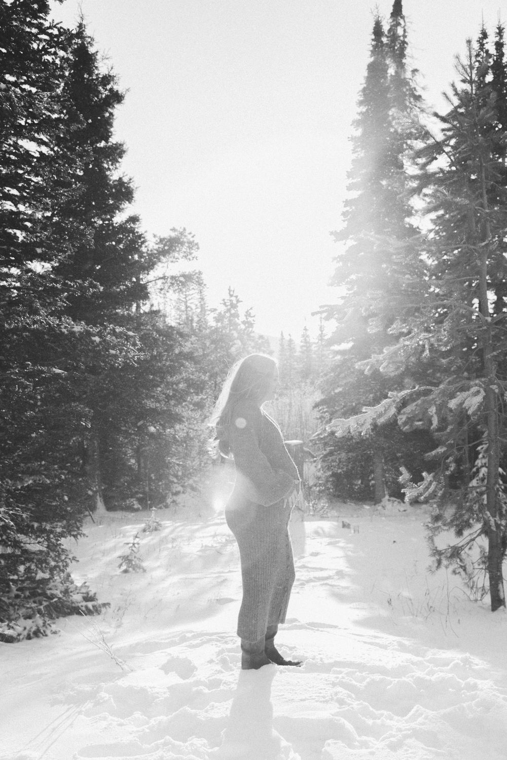 A silhouette of a person standing in a snow-covered forest with sunlight streaming through tall pine trees.