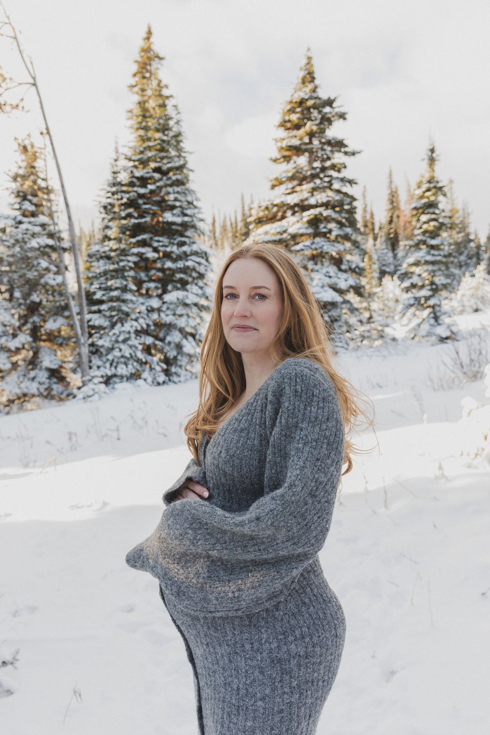 Person wearing a cozy gray sweater poses in a snow-covered mountain landscape with evergreen trees.