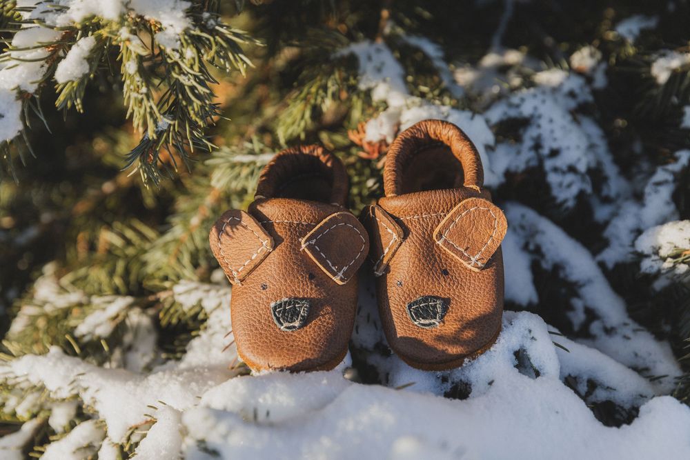 Small brown leather baby booties sitting on snow with evergreen branches in the background.