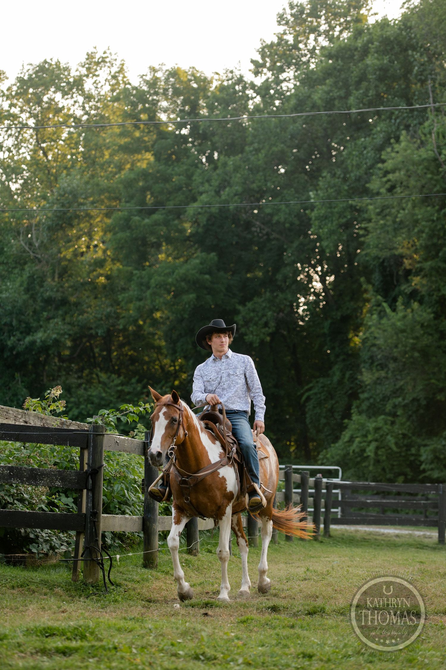 A paint horse and rider pose near a wooden fence on a summer evening.