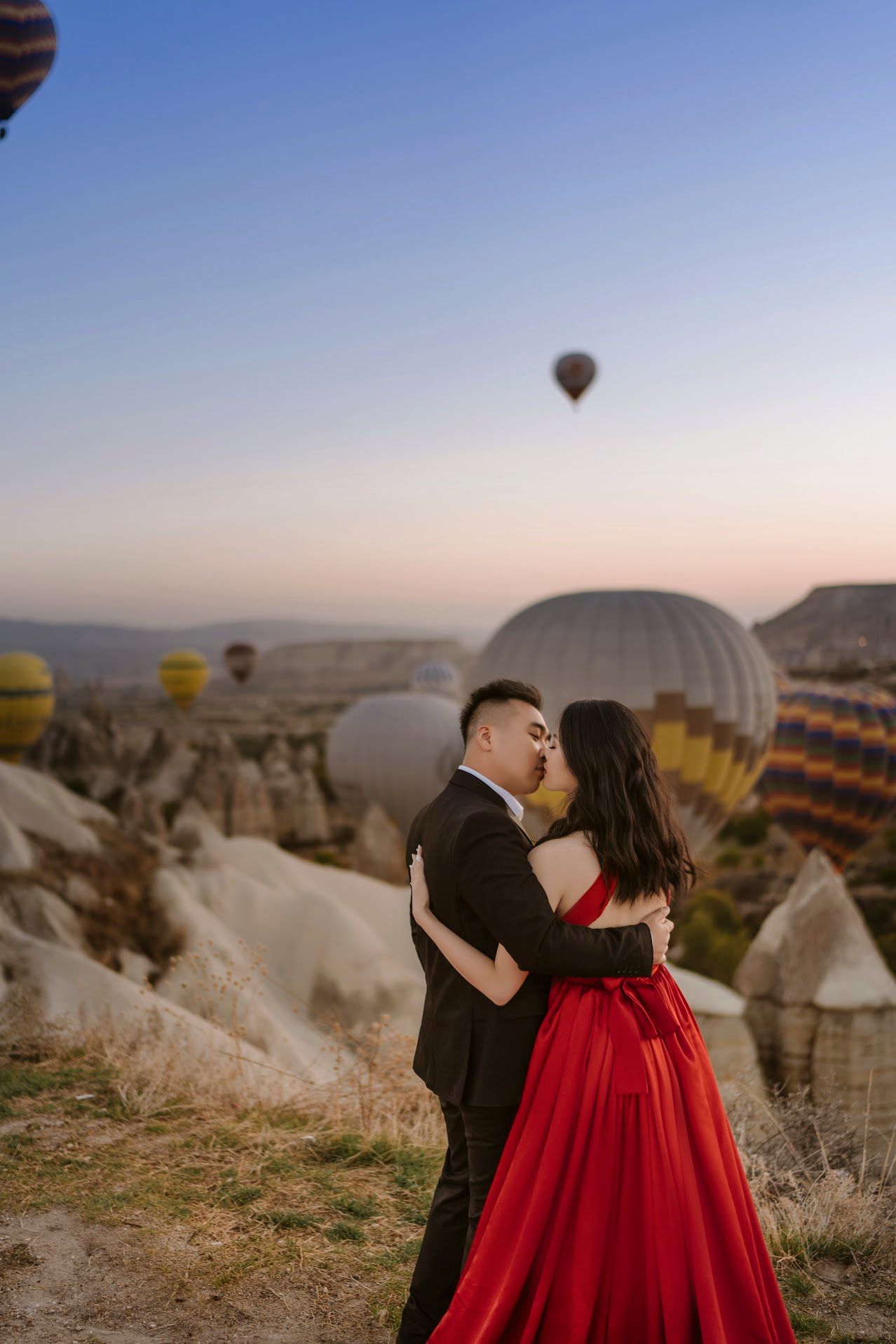 Couple in formal wear embraces on hillside while hot air balloons float over Cappadocia's distinctive landscape at sunrise.