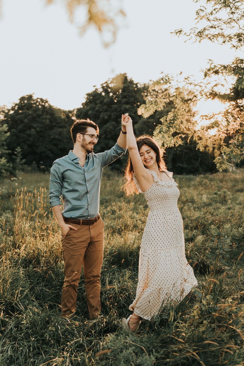 A couple dances in a sunlit meadow at golden hour, wearing casual attire as warm light filters through the trees.