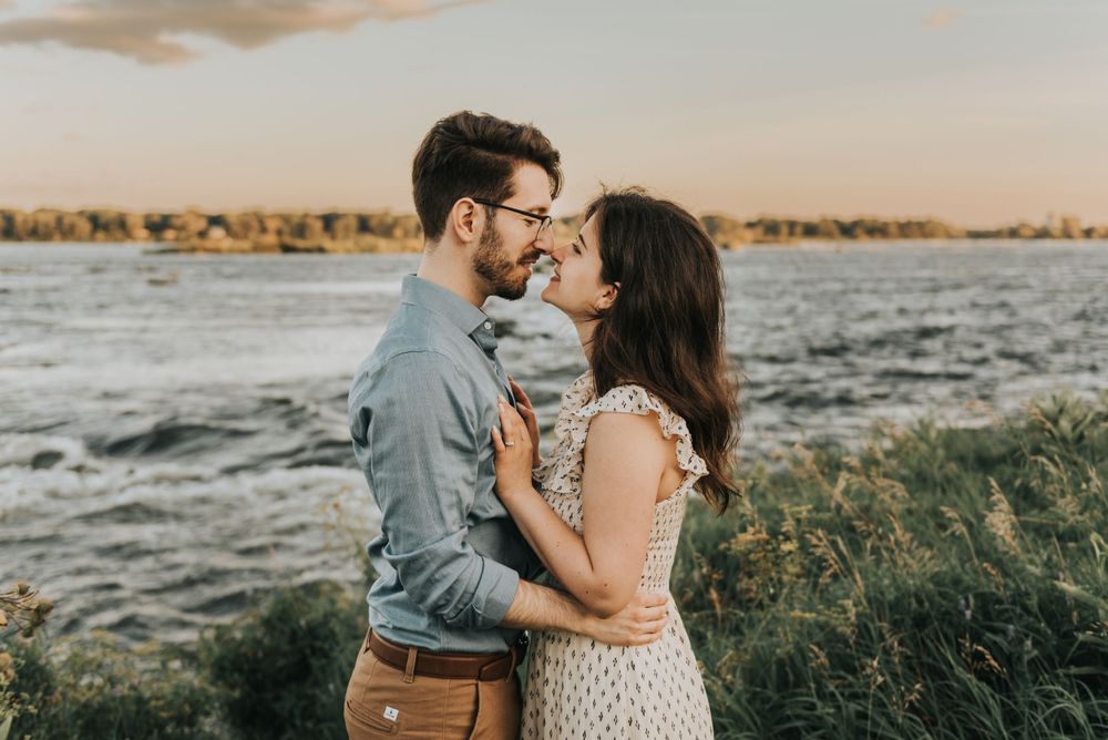 A romantic couple embraces by a waterfront at sunset with golden light illuminating the natural landscape.