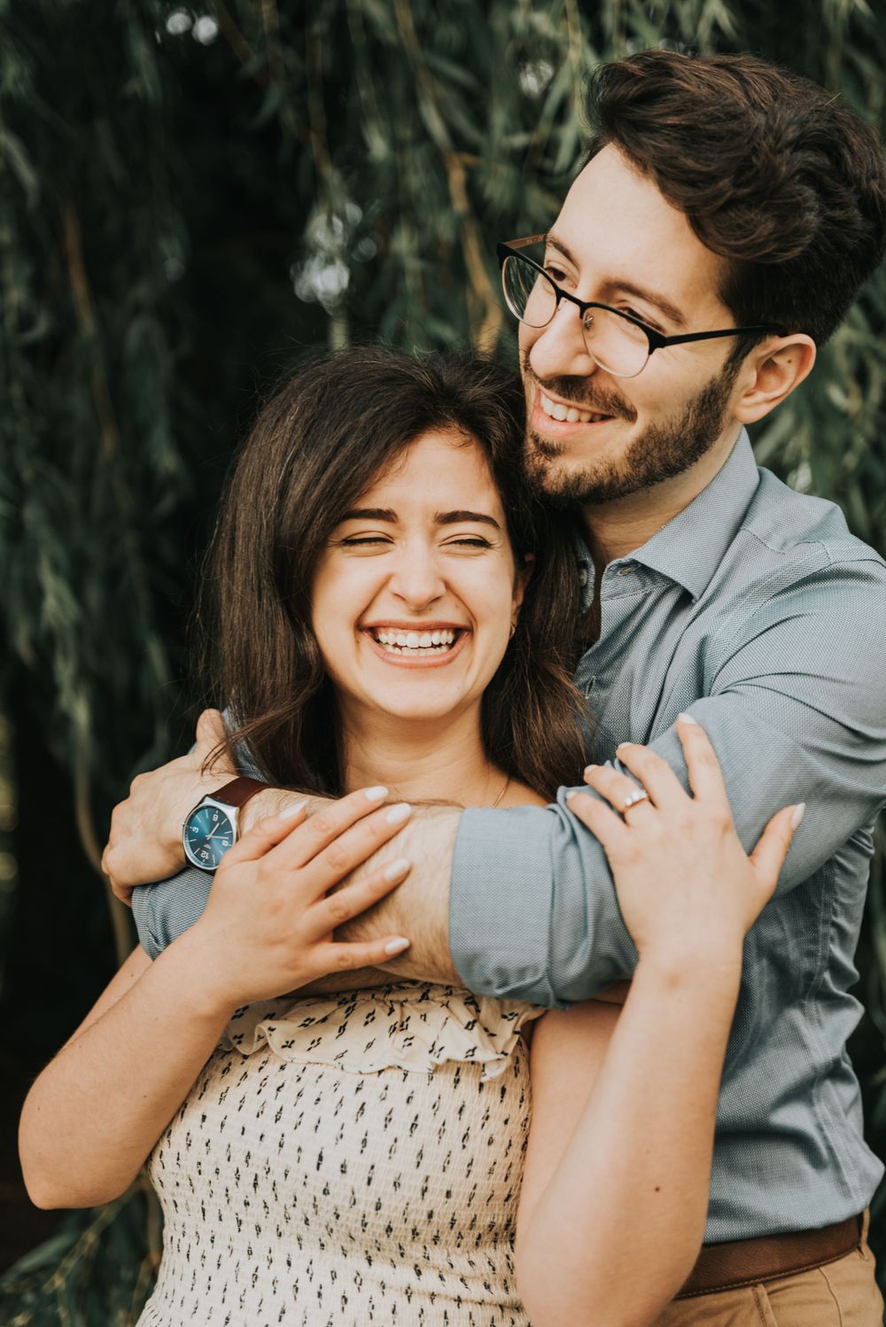A happy couple embraces and laughs together outdoors against a natural foliage backdrop.