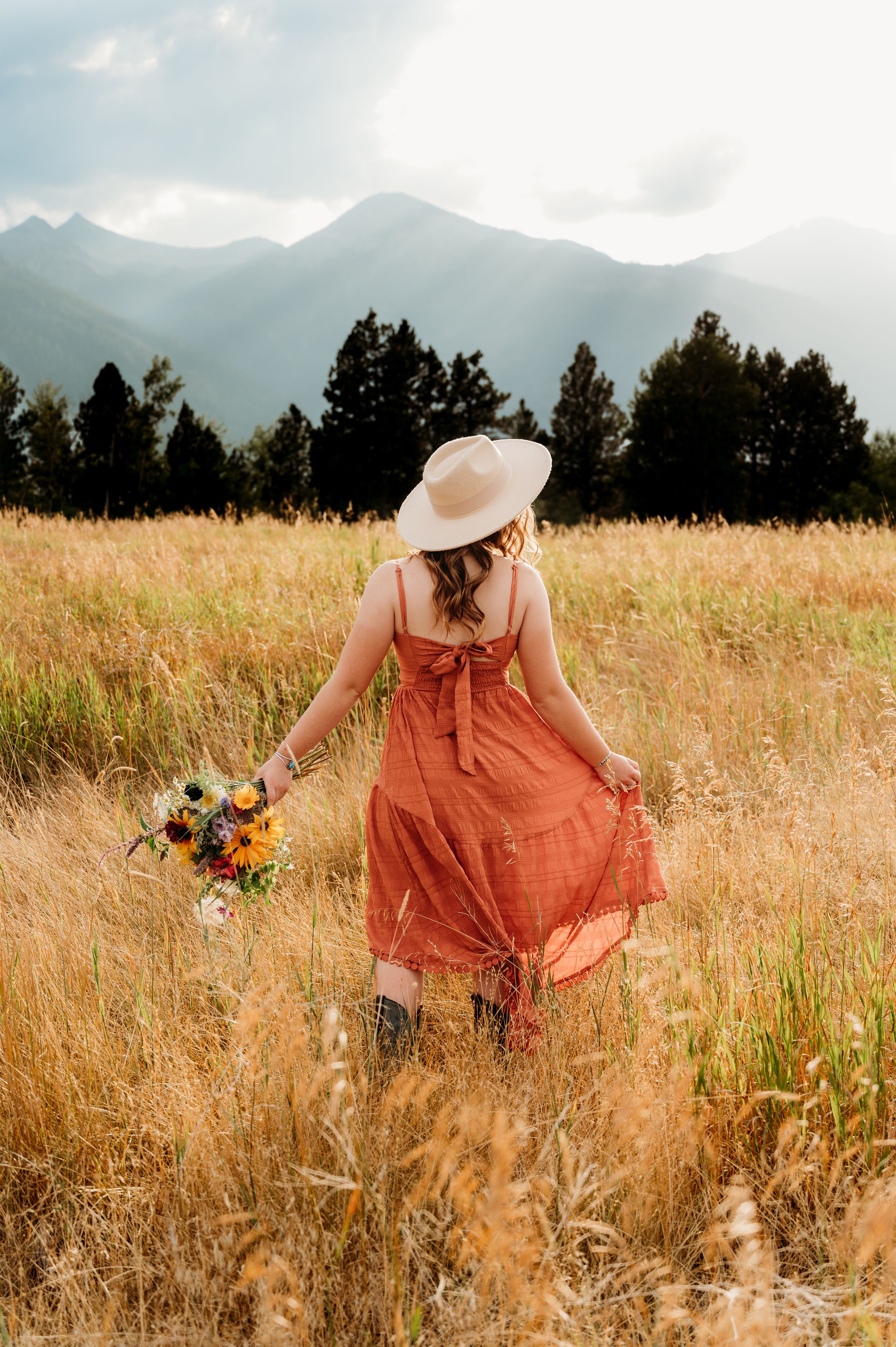 Person in orange dress holding wildflowers walks through golden mountain meadow. Wallowa Lake, Joseph, Oregon