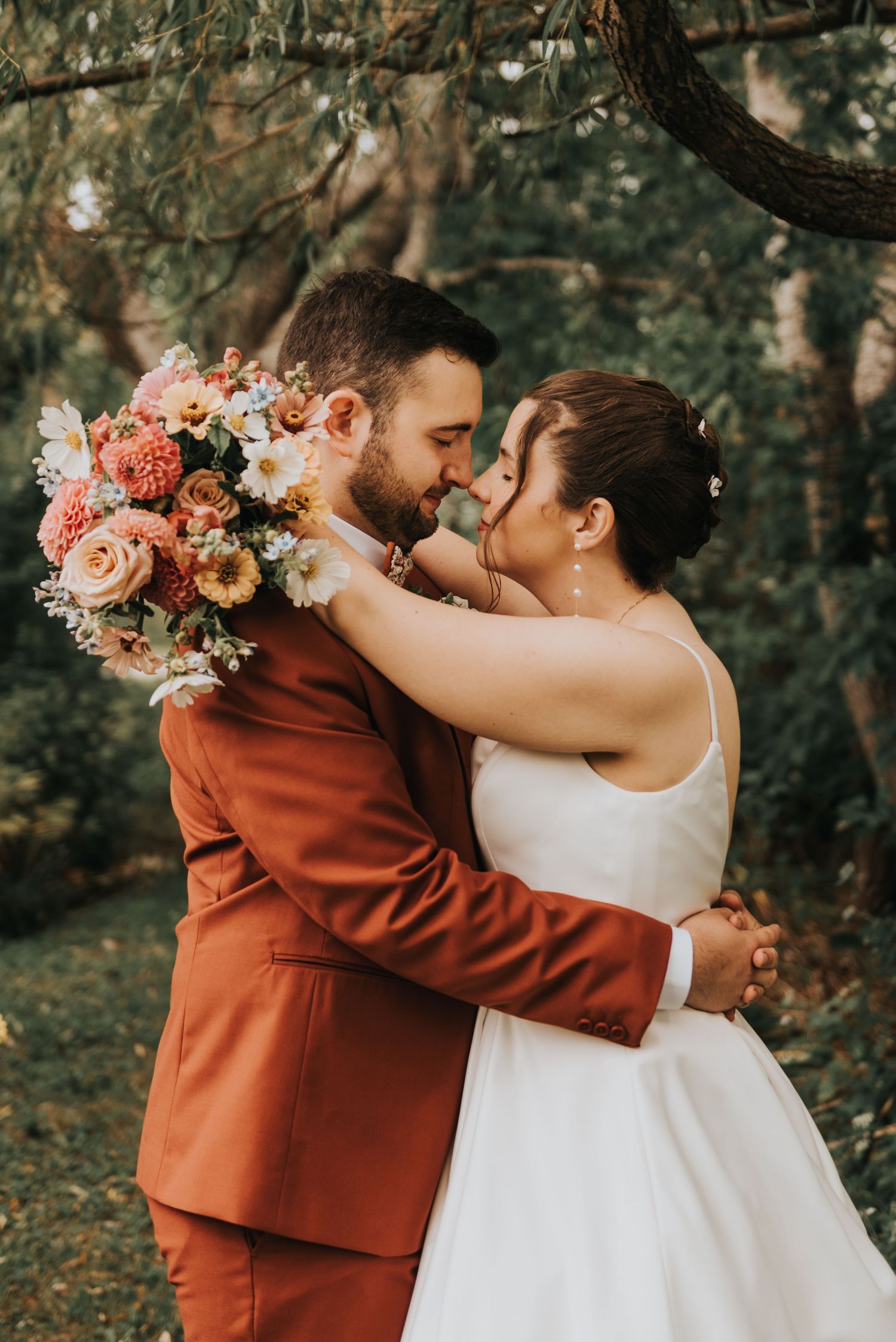 A couple in wedding attire share an intimate moment outdoors while holding a beautiful peach and blue floral bouquet.