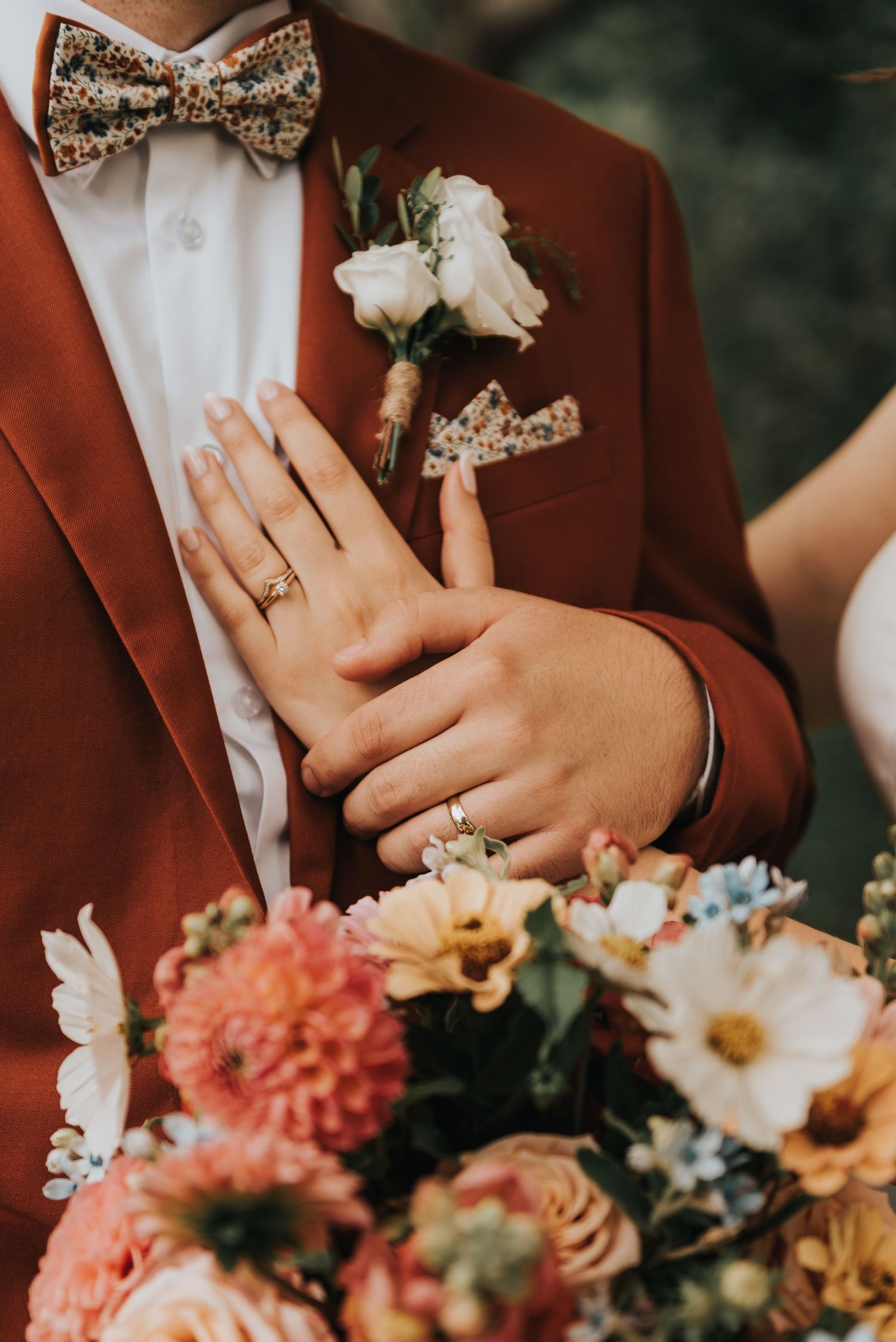 Close-up of maroon suit jacket with floral bow tie, white boutonniere, and hand with wedding ring against colorful bouquet.