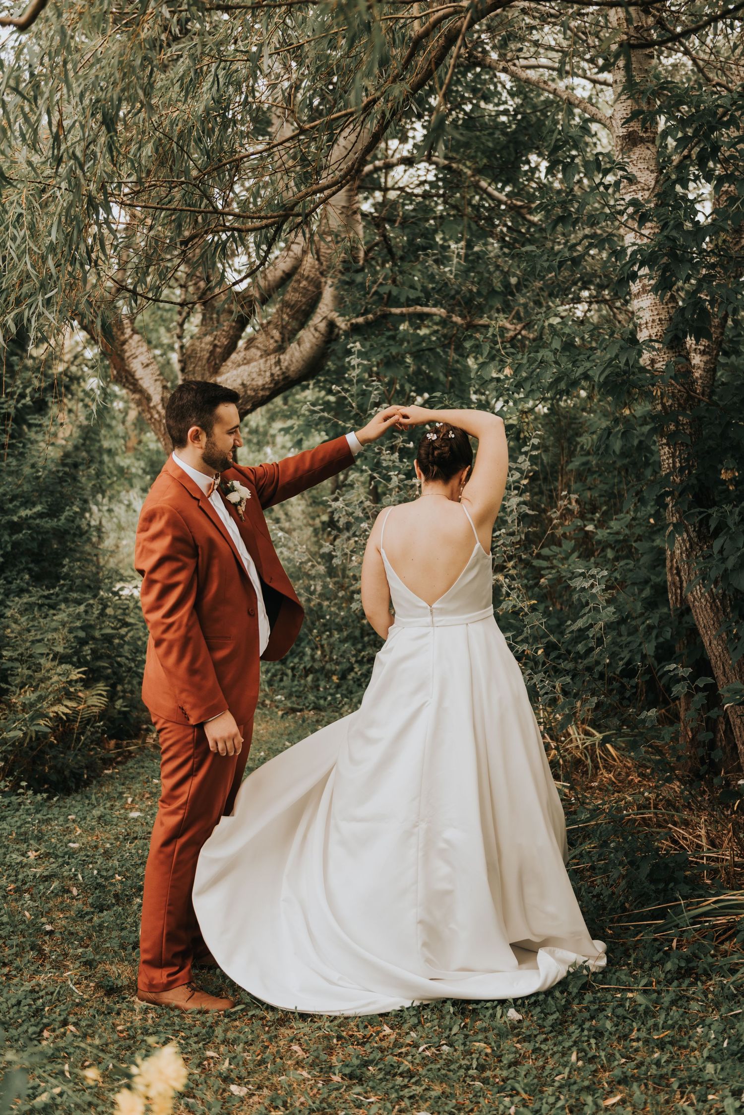 Couple in a rustic forest wedding scene with bride in white gown and groom in rust-colored suit.