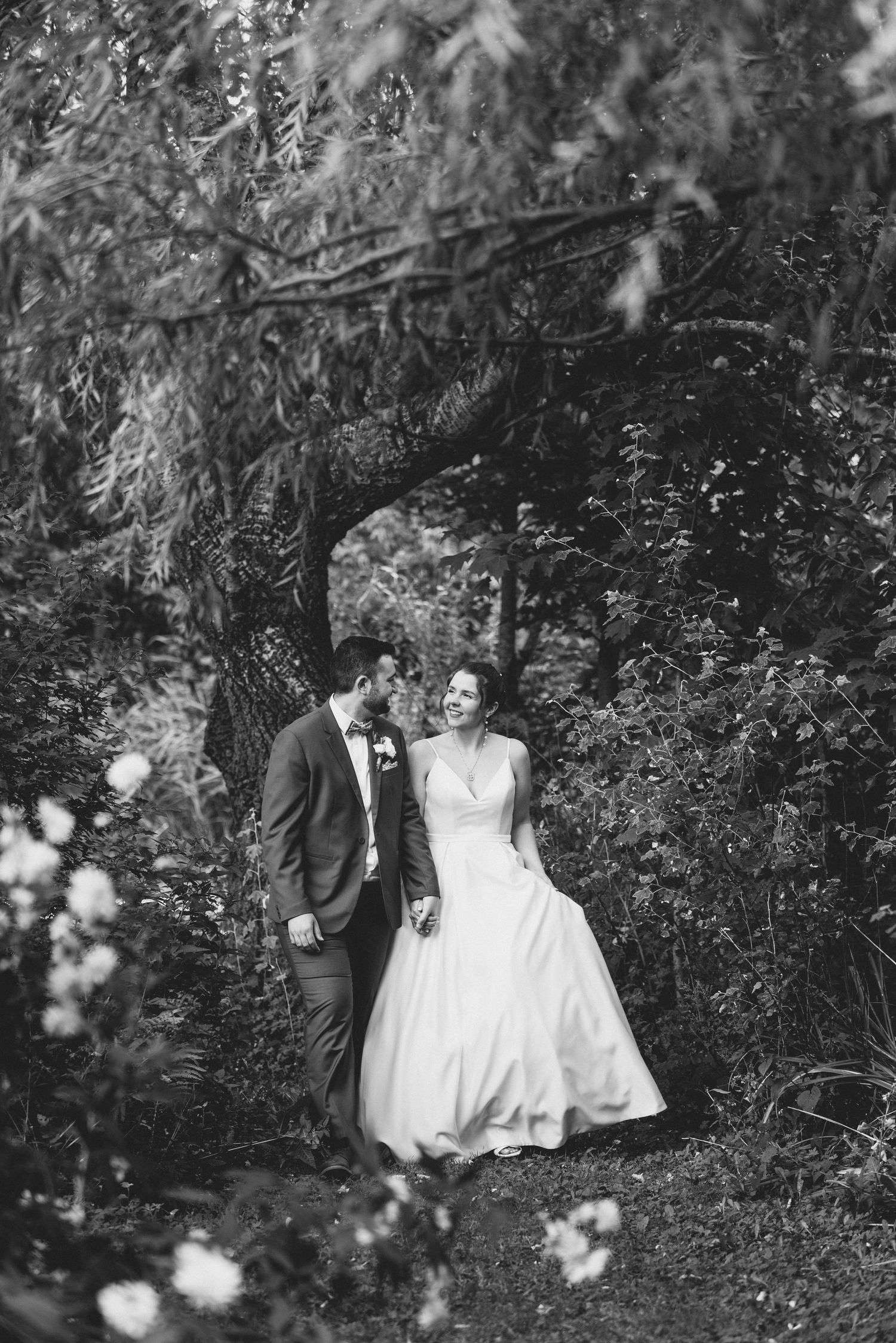 A romantic black and white wedding photograph captured in a lush garden setting with blooming flowers in the foreground.