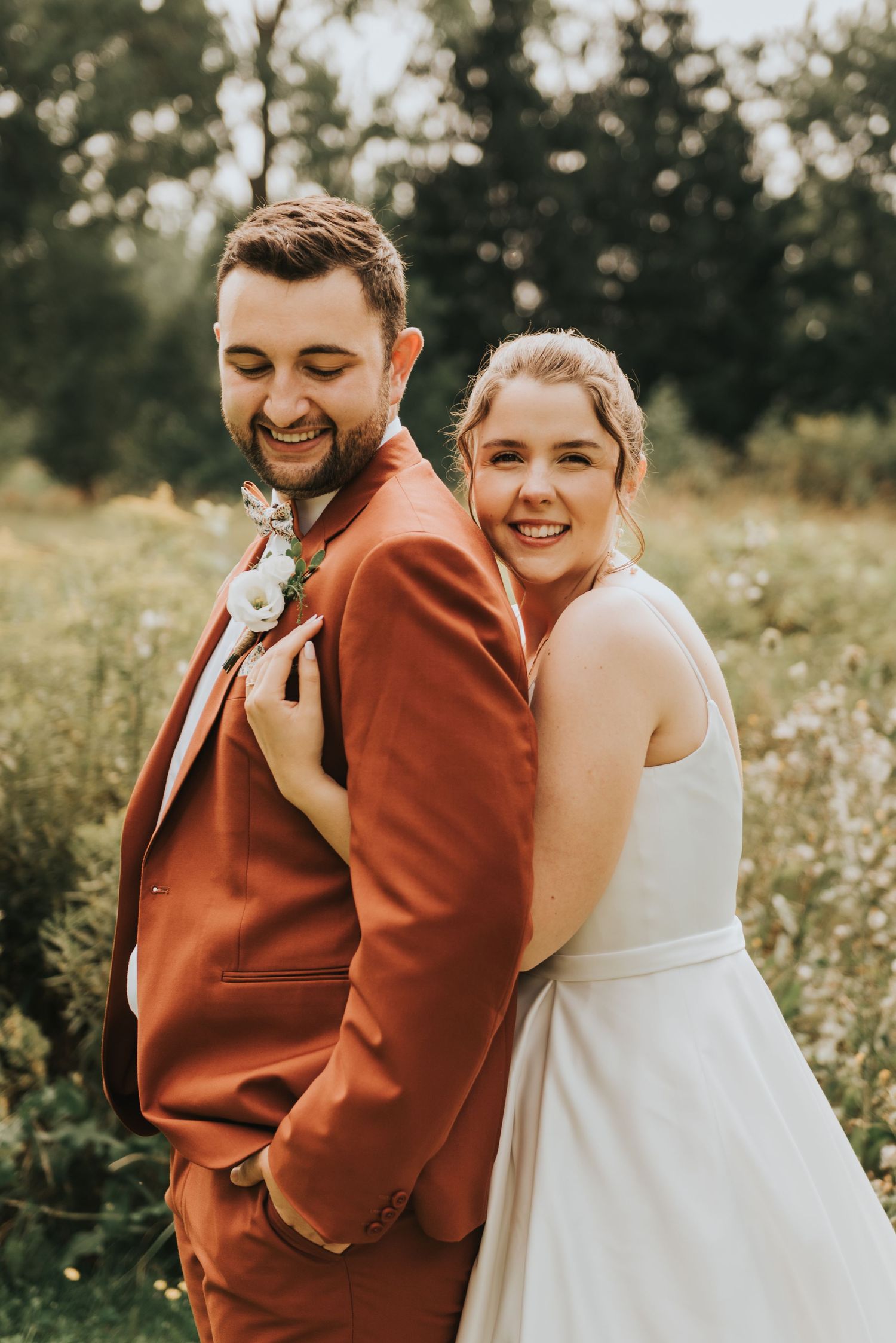 A happy couple in a wildflower field, with the groom in a rusty orange suit and bride in a white wedding dress.