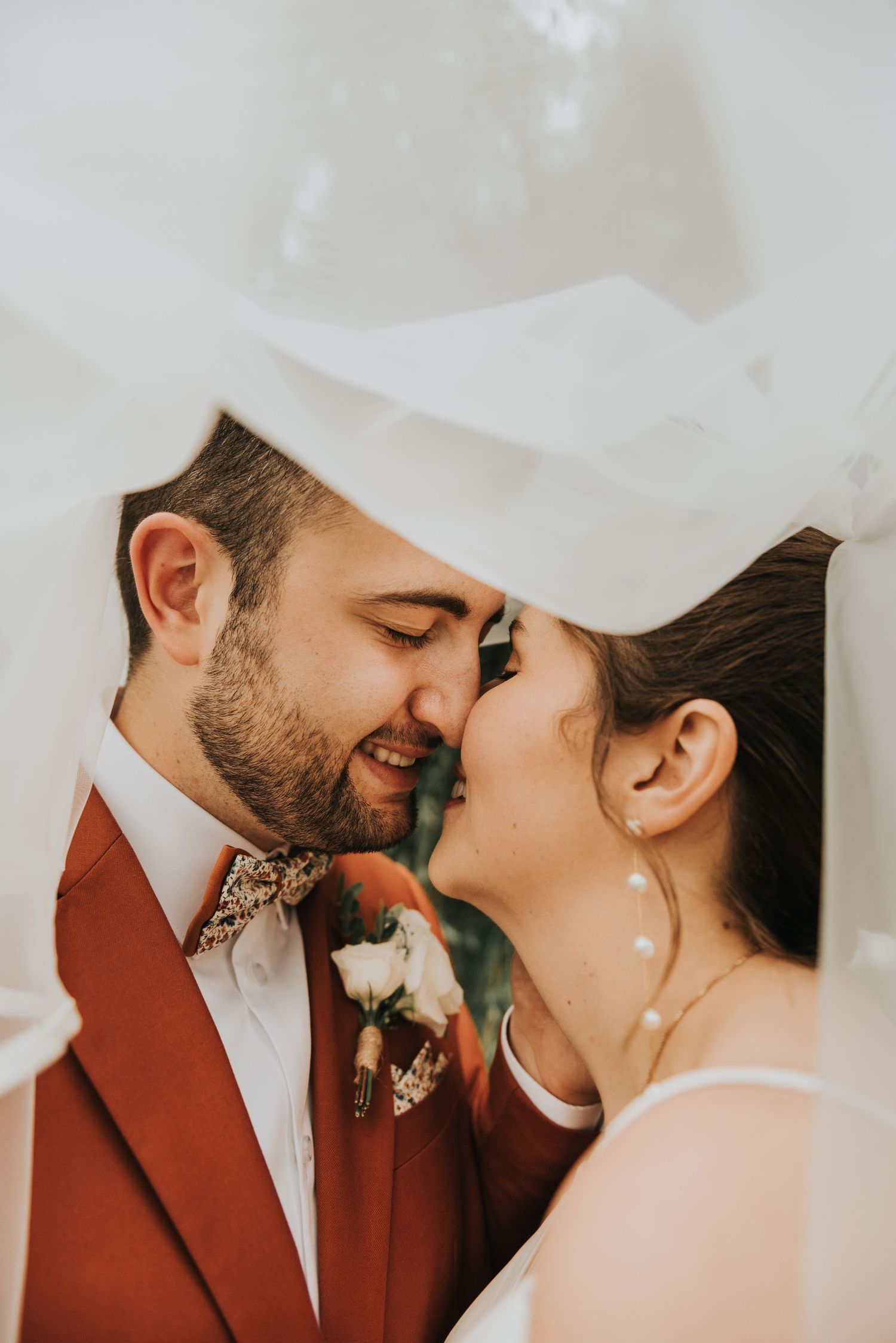 A romantic wedding moment captured between newlyweds under a flowing white veil with a burgundy suit and floral boutonniere.