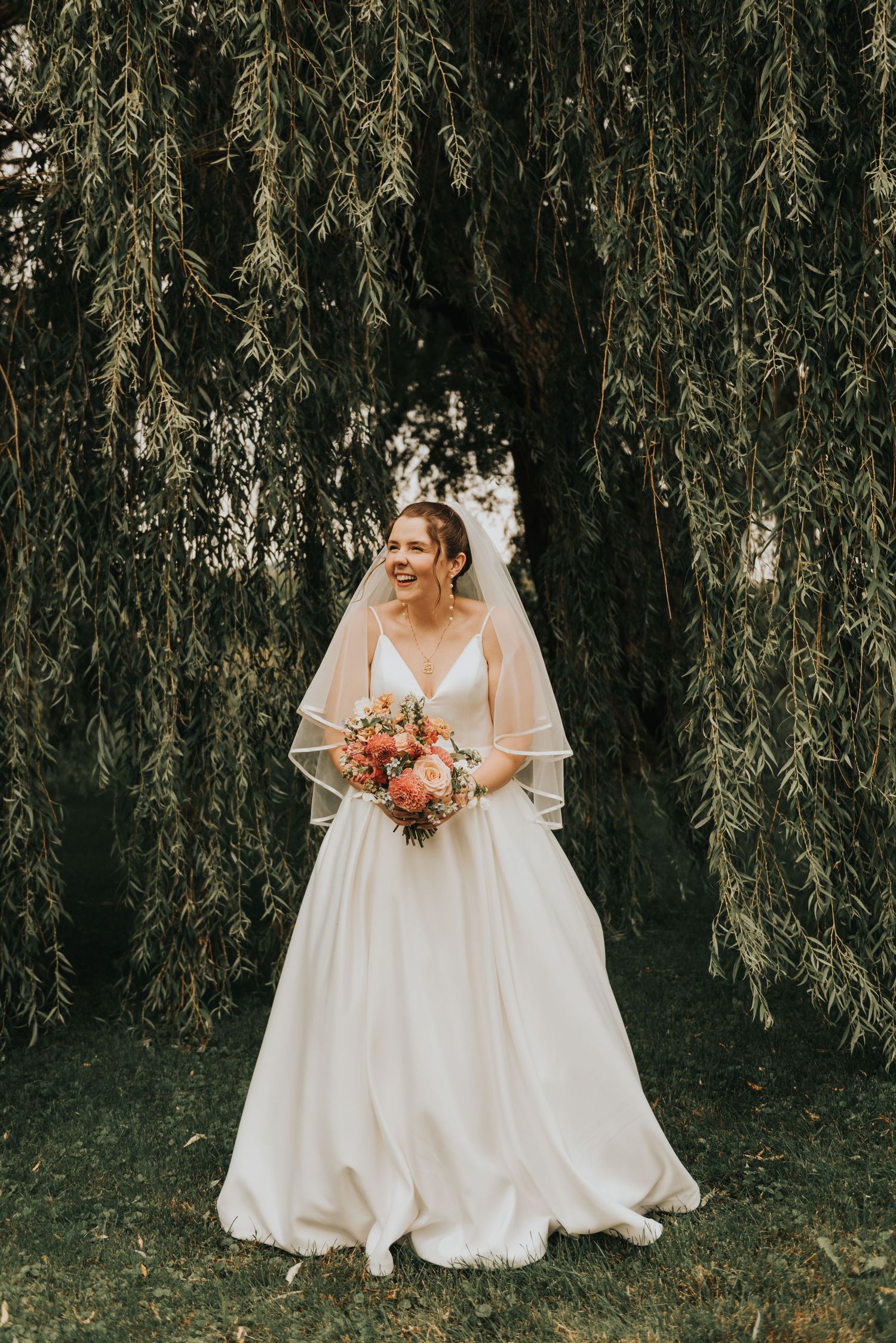A person in an elegant white wedding gown holds a coral bouquet while standing beneath a dramatic weeping willow tree.