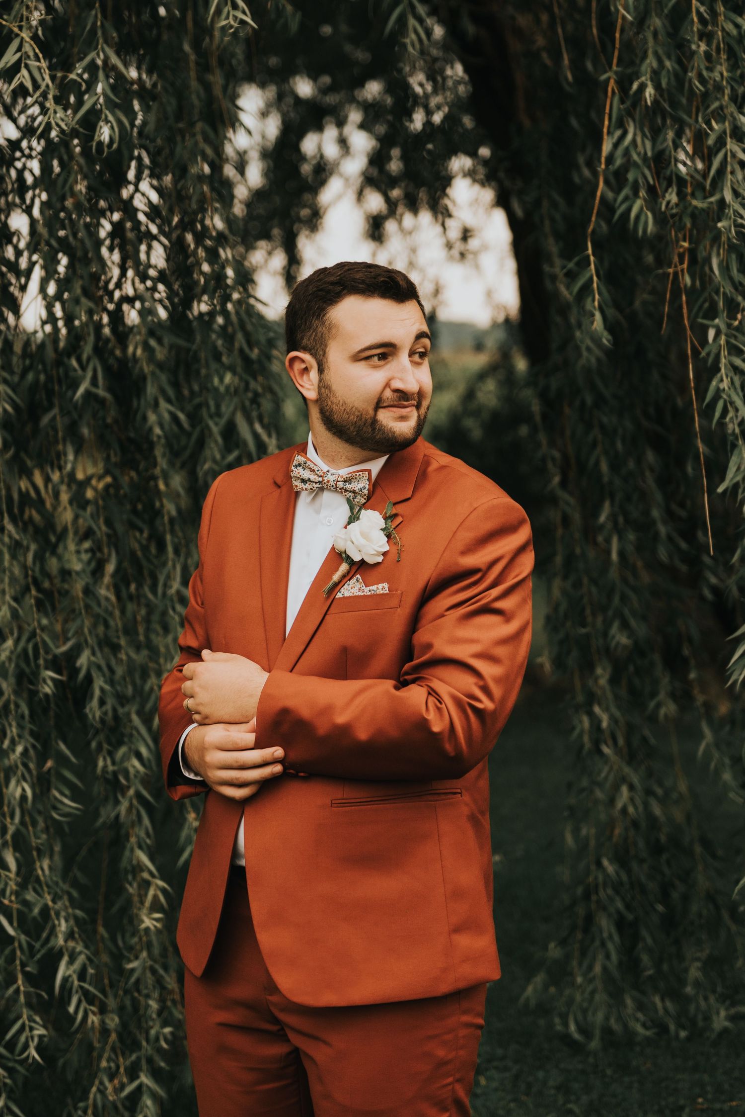 A groom in a striking burnt orange suit and bow tie stands among hanging willow tree branches during a wedding photoshoot.