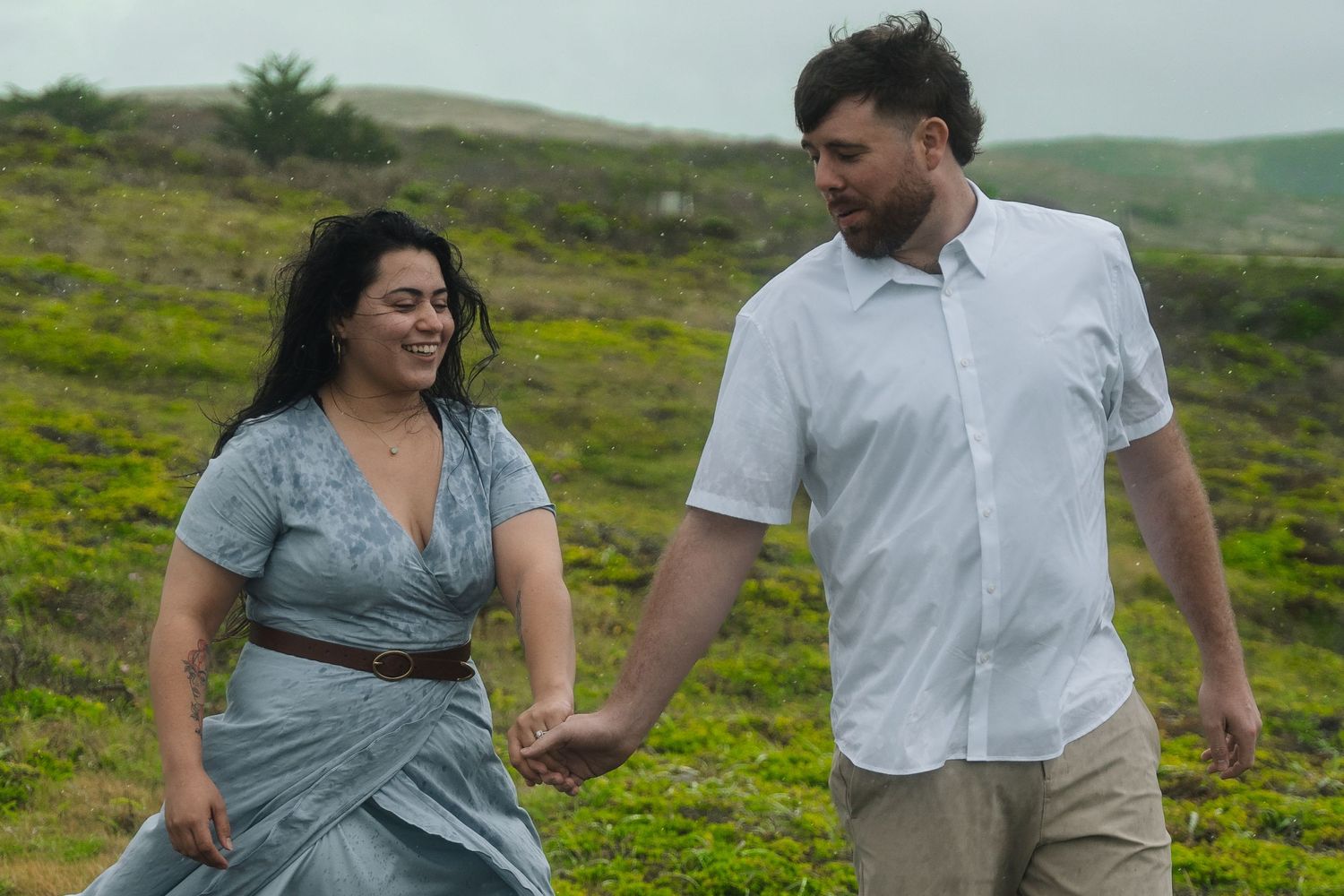 Two people hold hands while walking together through a green hillside meadow on a cloudy day.