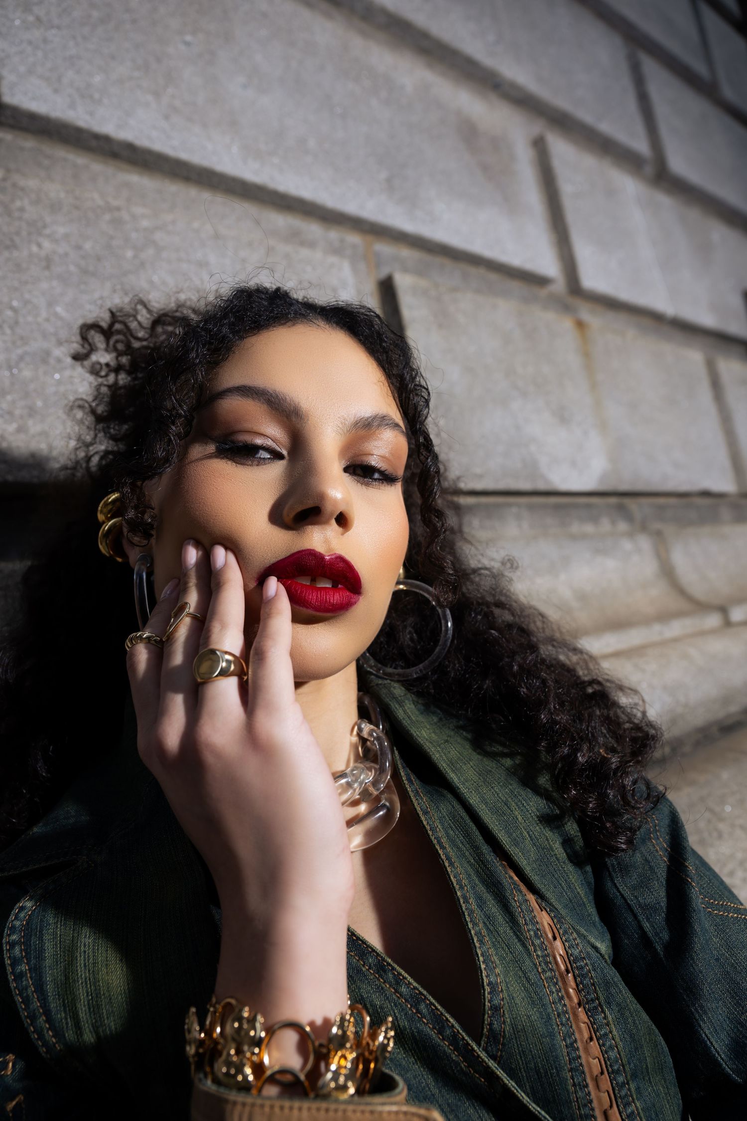 Dramatic portrait with bold red lipstick and gold accessories against stone wall background.