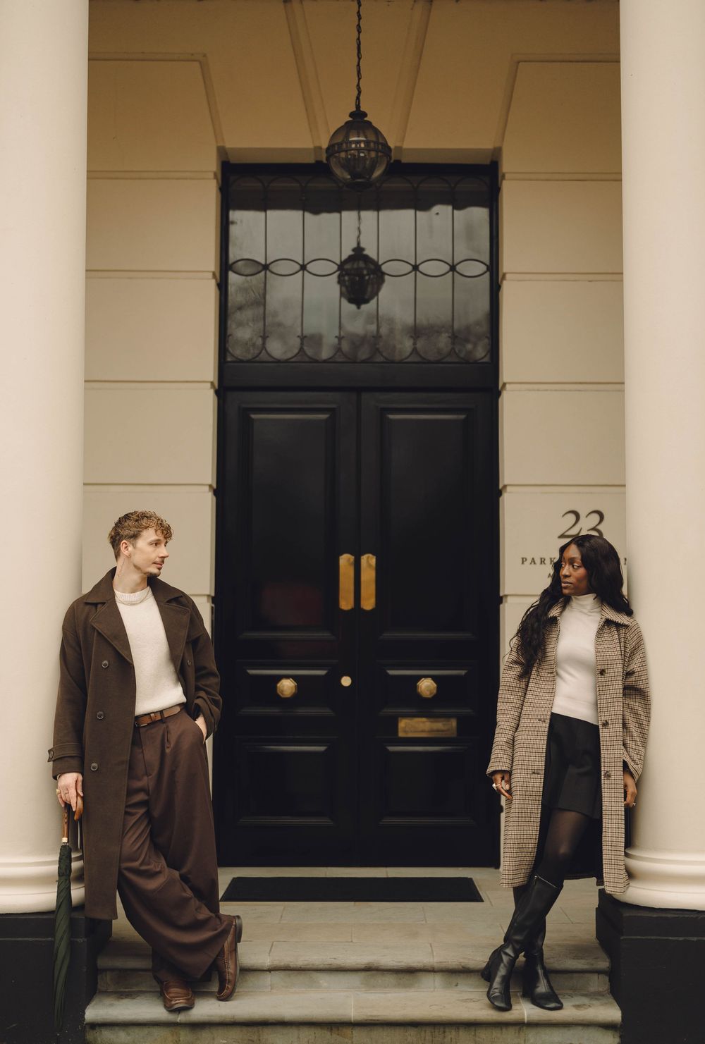 Two people in stylish winter coats pose in front of a classic black door with ornate details in London.