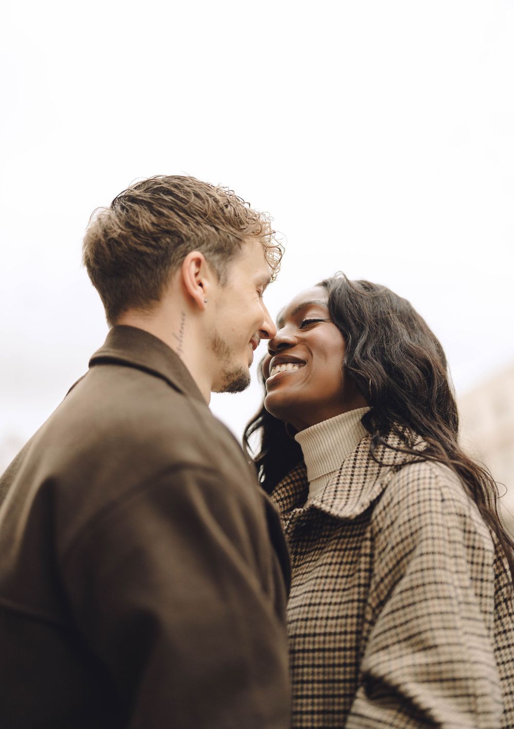 A couple in winter coats sharing an intimate moment and laughing together against a bright background.