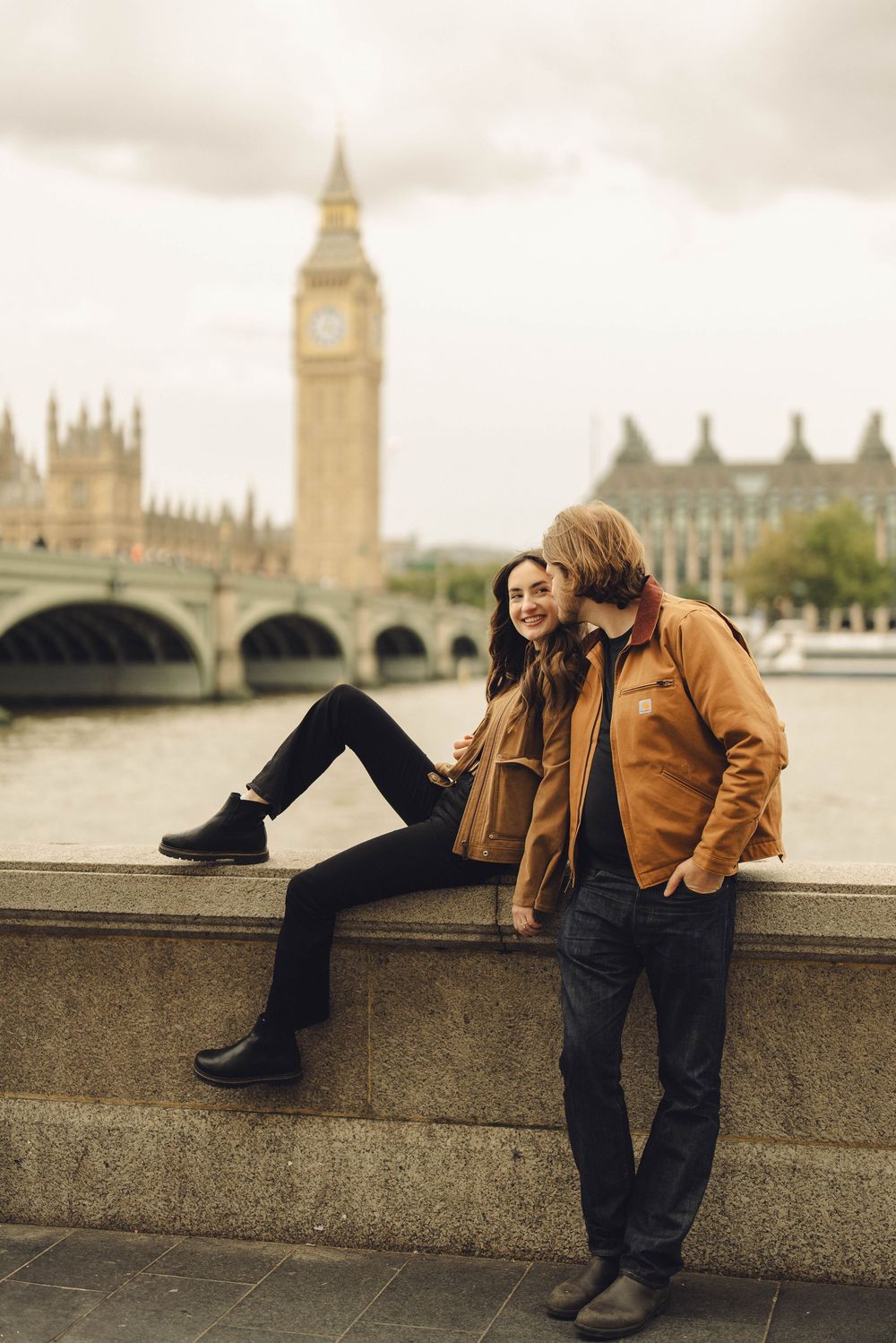 A couple in brown jackets sits along the Thames River with Big Ben and Westminster Bridge in view.