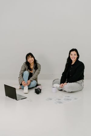 Two people sit on the floor in a bright minimalist setting with a laptop and camera equipment between them.
