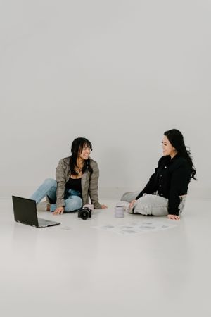 Two women sitting on the floor having casual conversation with laptop and coffee in minimalist white setting.