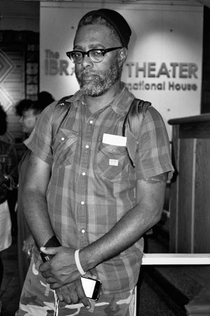 A black and white photo of Arthur Jafa wearing glasses and a plaid shirt standing in front of a theater sign at the Black Star Film Festival
