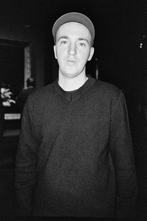 A black and white portrait of artist KAWS wearing a dark v-neck sweater and baseball cap against a dark background.