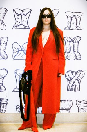 Aubrie Costello in bold red pantsuit and sunglasses stands against wall with fashion sketch artwork background.