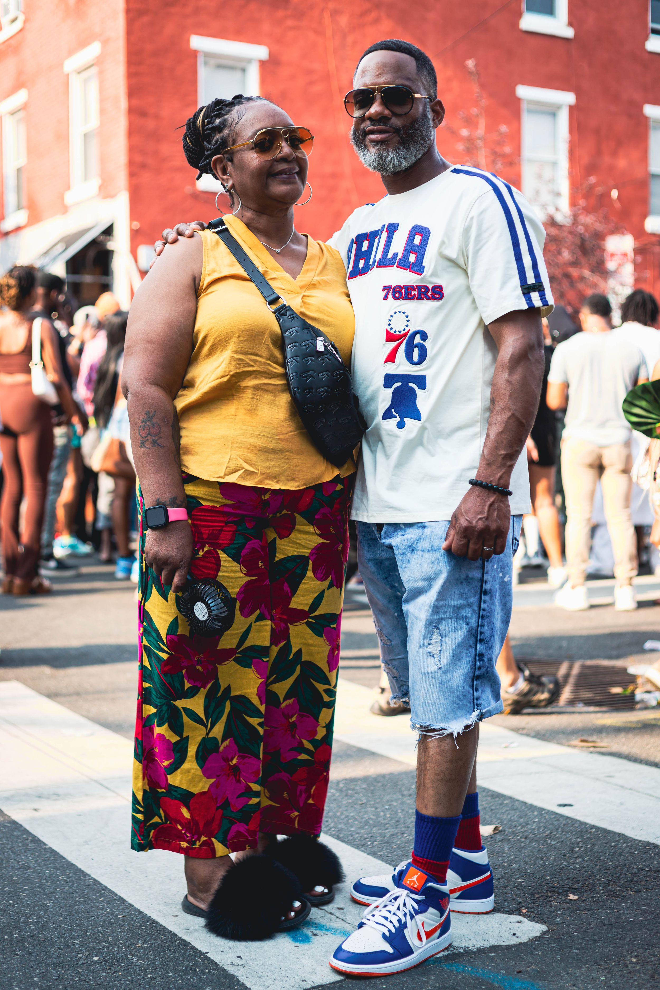 A couple wearing colorful summer outfits pose together at an outdoor event, with stylish sneakers and vintage-inspired clothing at ODUNDE
