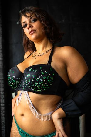 Portrait photo with dramatic lighting showcasing a black bra embellished with green gems, silver fringe and dark background.
