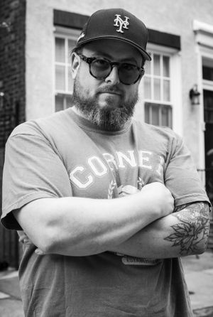 Black and white portrait of Iron Solomon wearing glasses, NY Mets cap and Coney t-shirt with visible arm tattoos.