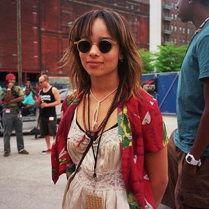 Zoe Kravitz wearing a red floral shirt, sunglasses and layered necklaces stands outdoors in an urban setting.