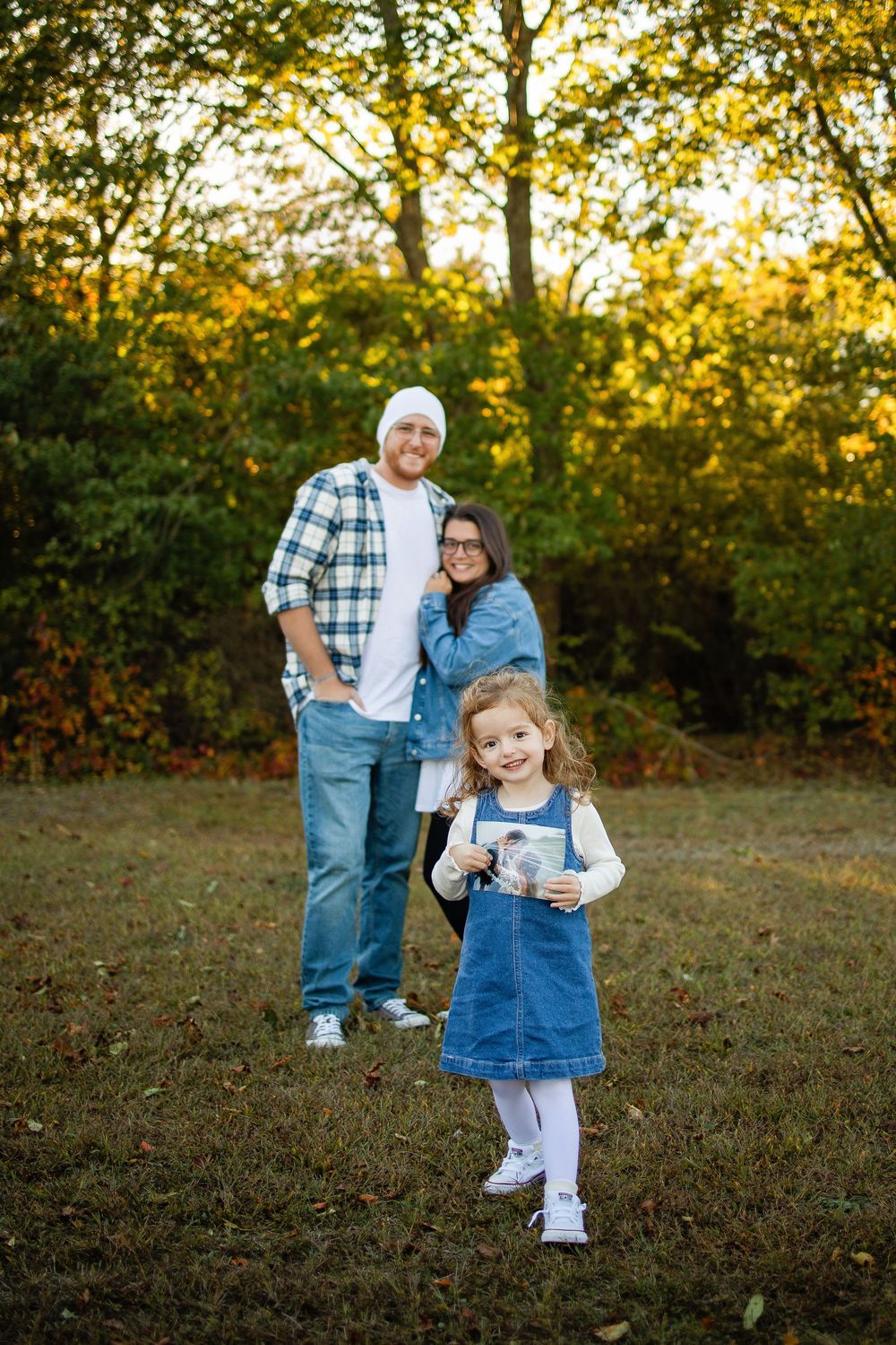 A family in casual attire poses together on autumn grass with golden sunlight filtering through trees in the background.