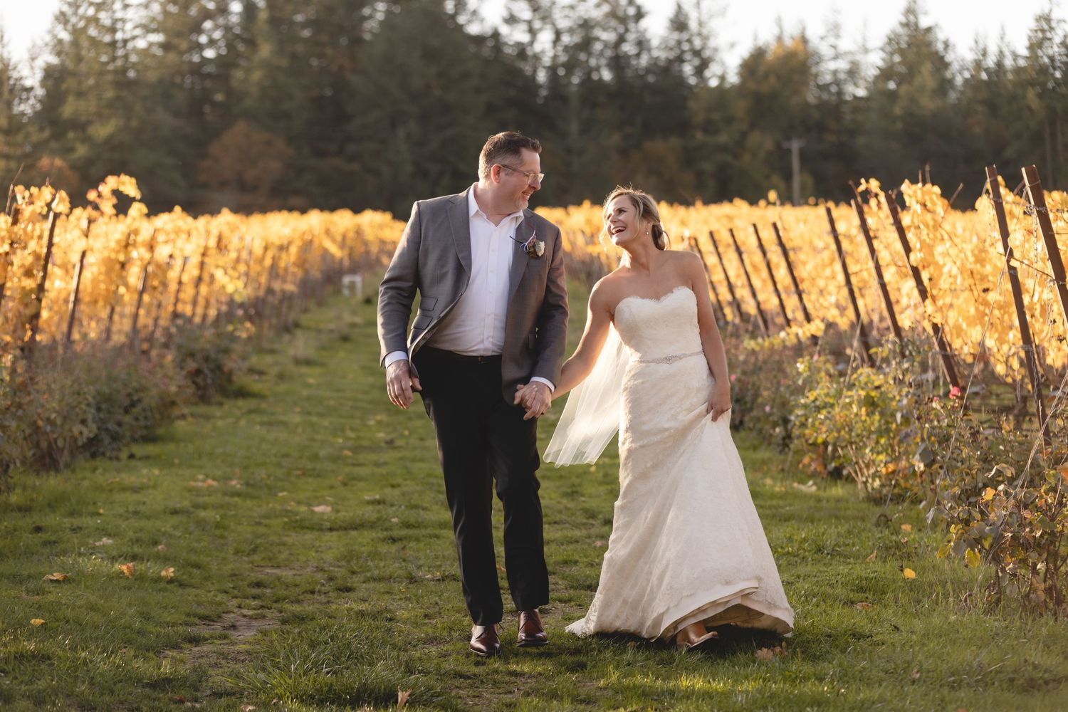 A couple walks hand in hand through a golden vineyard at sunset, with the bride's white dress flowing in the evening light.