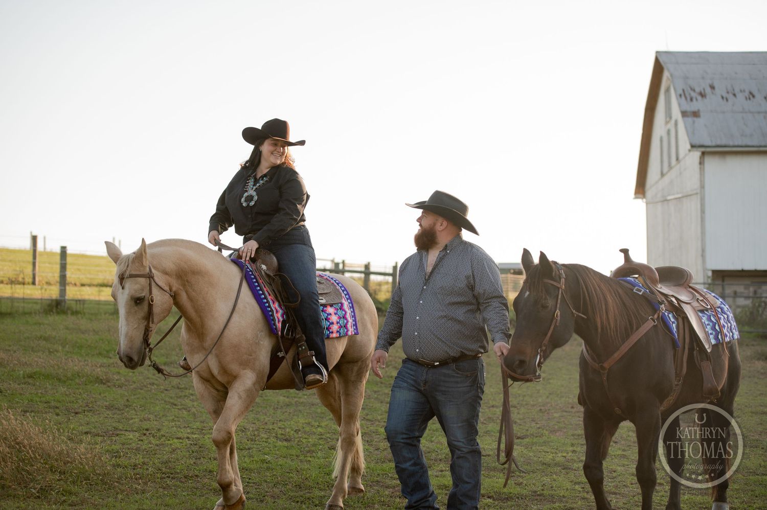 Two horses with western tack and riders stand near a white barn at sunset.