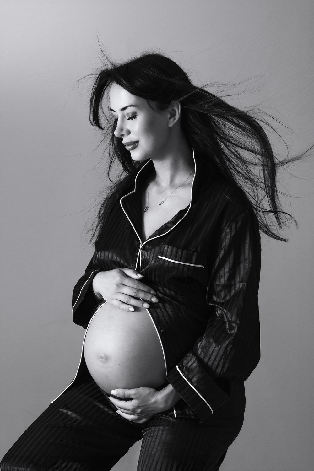 Black and white maternity photo series showing pregnant woman in dark sweater with flowing hair against grey backdrop.