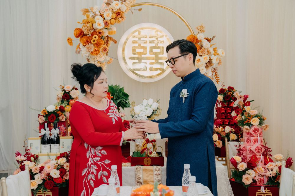 Woman in red ao dai presents gift to groom in blue ao dai at traditional Vietnamese wedding.