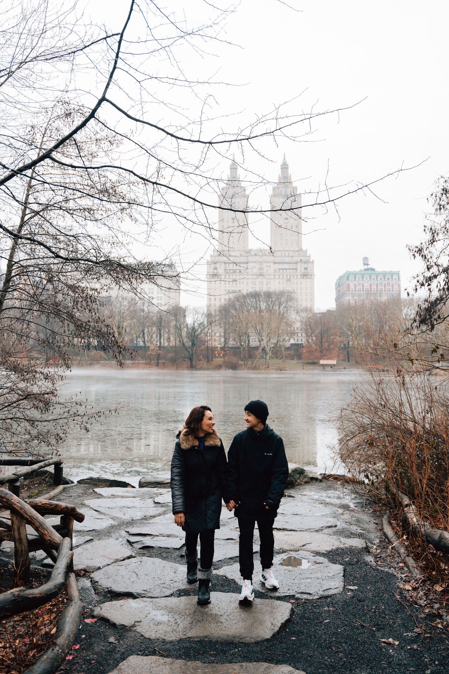 Couple walking along Central Park lake with foggy NYC skyline in background.