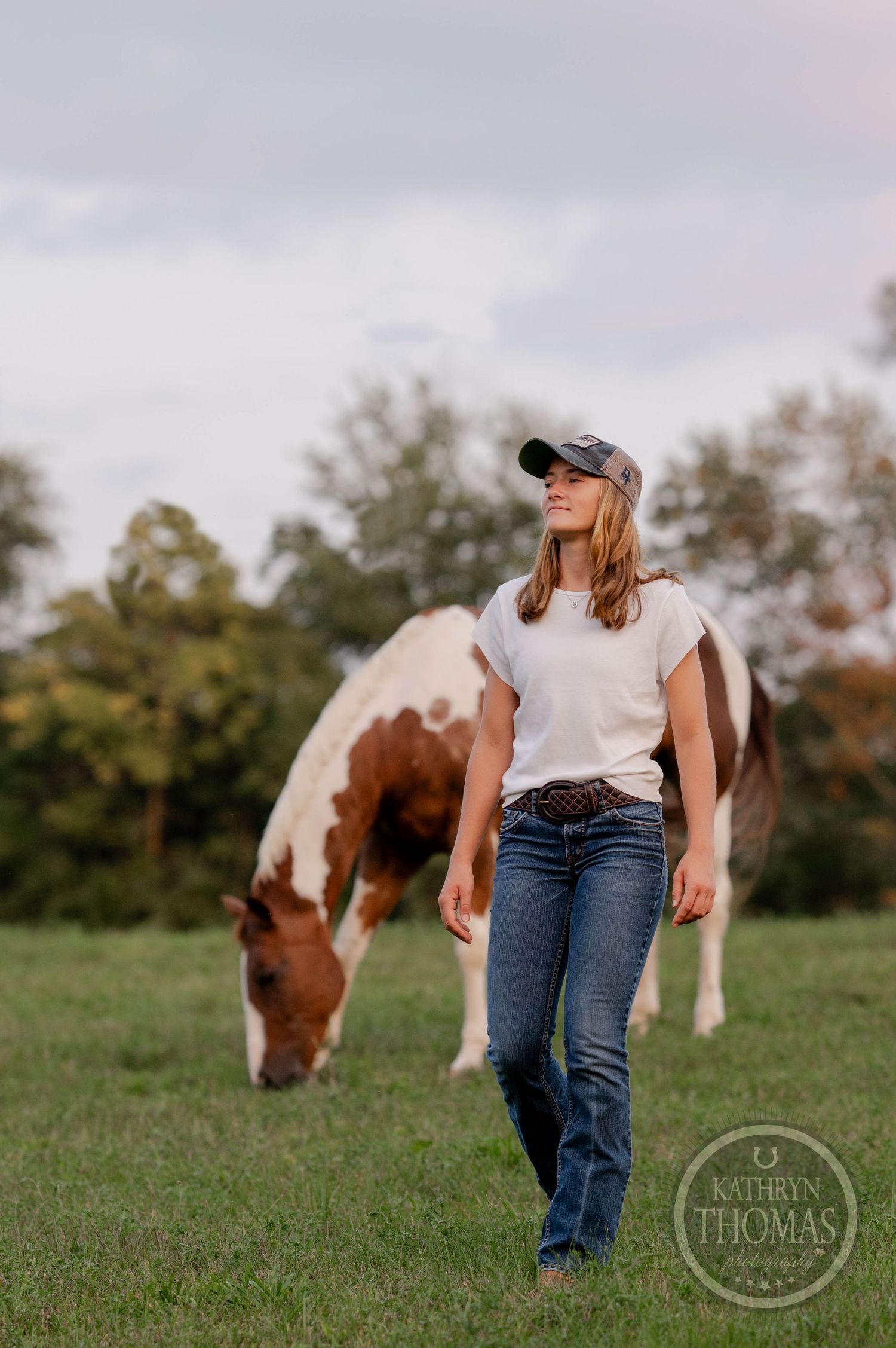 A paint horse grazes peacefully in a field while someone in jeans and a baseball cap walks by.
