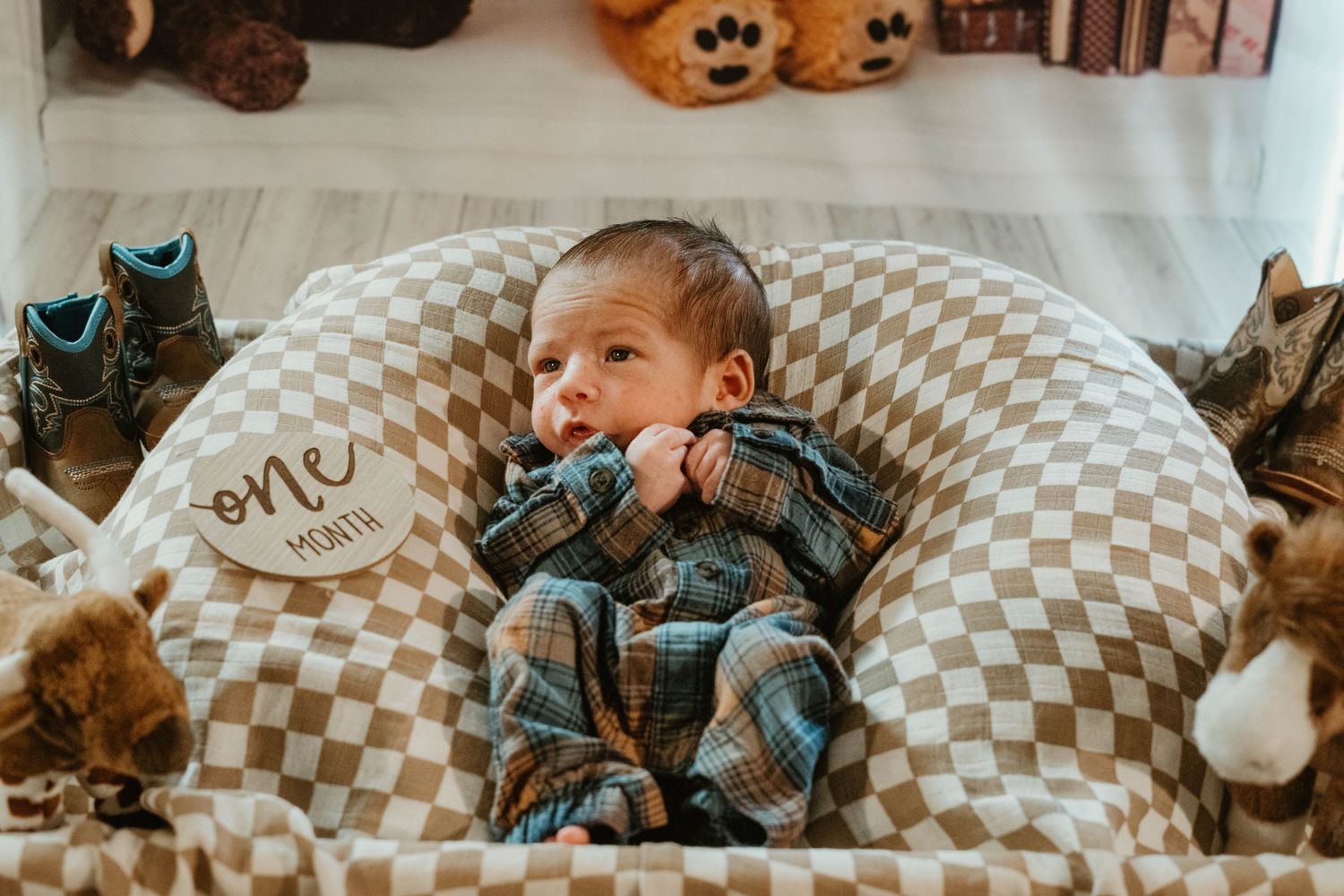 A baby rests on a cozy checkered pillow wearing a plaid flannel outfit while surrounded by stuffed animals.