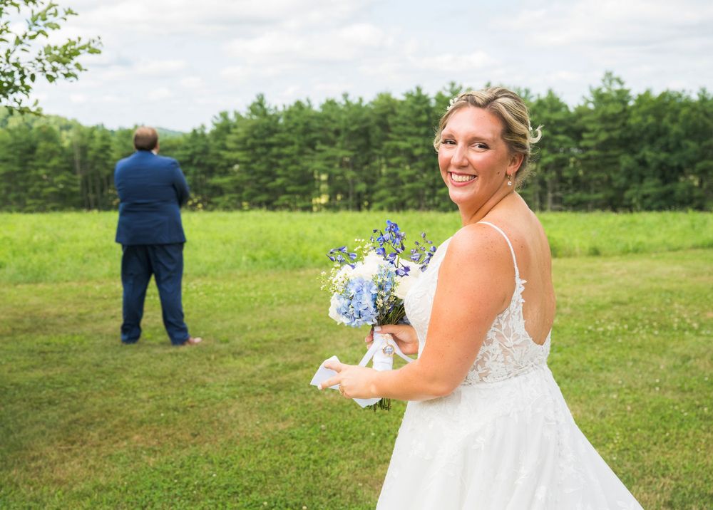 A couple in wedding attire poses in a lush green meadow surrounded by trees in summer.