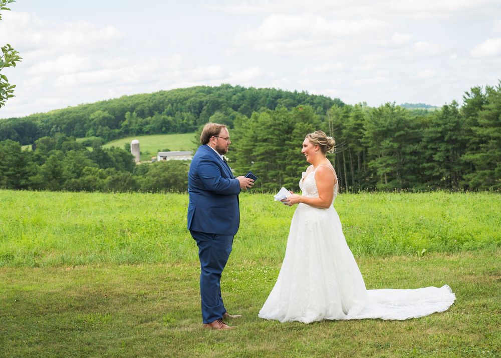Wedding couple embraces while standing in a scenic rural field with pine forest in the background.